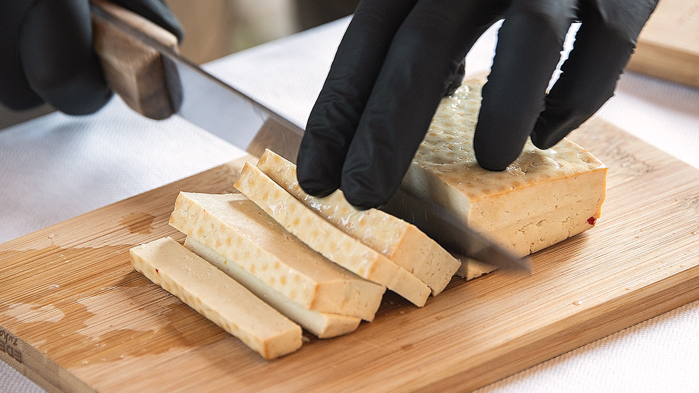 Hands in black gloves slicing firm tofu on a wooden cutting board.