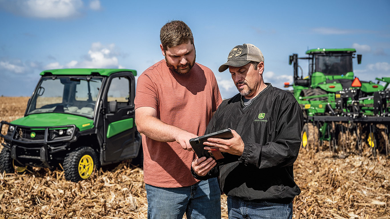 Two people using tablet in field; green farming equipment behind