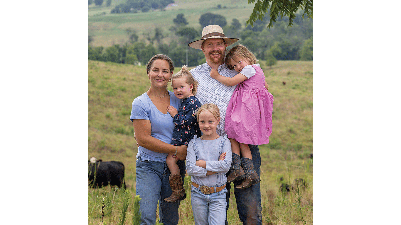 Two adults and three children standing together in a grassy pasture with cattle.