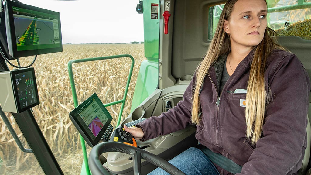 In-cab photo of a young farmer operating a John&nbsp;Deere S7 Combine in corn. You can see before them a G5<sup>Plus</sup> Display, a New Generation Primary Corner Display and a G5<sup>Plus</sup> Extended Monitor.