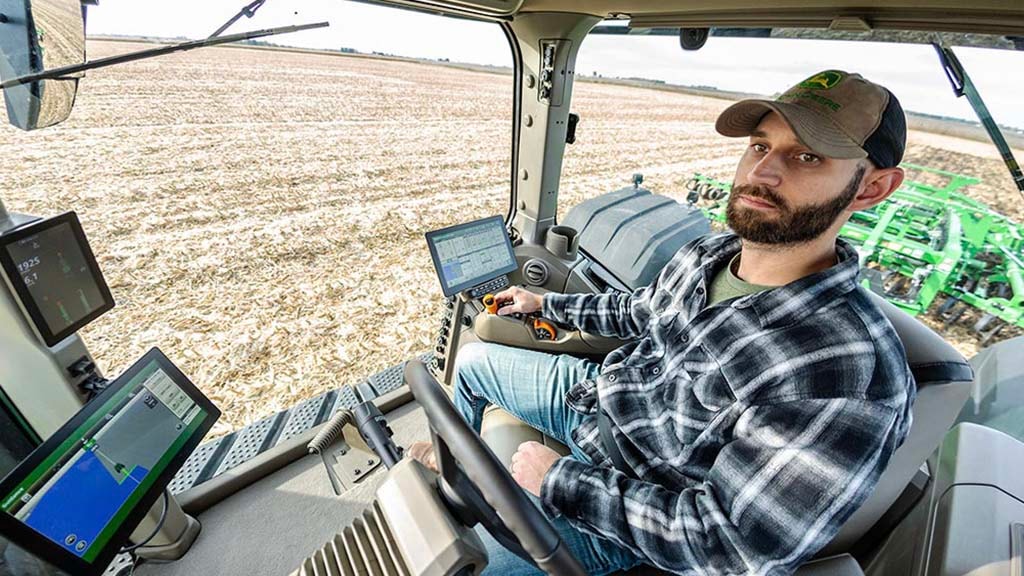 In-cab photo of a young farmer operating a John&nbsp;Deere 9RX tractor. You can see before him a G5Plus Display, a G5<sup>Plus</sup> Extended Monitor and a large right side window.