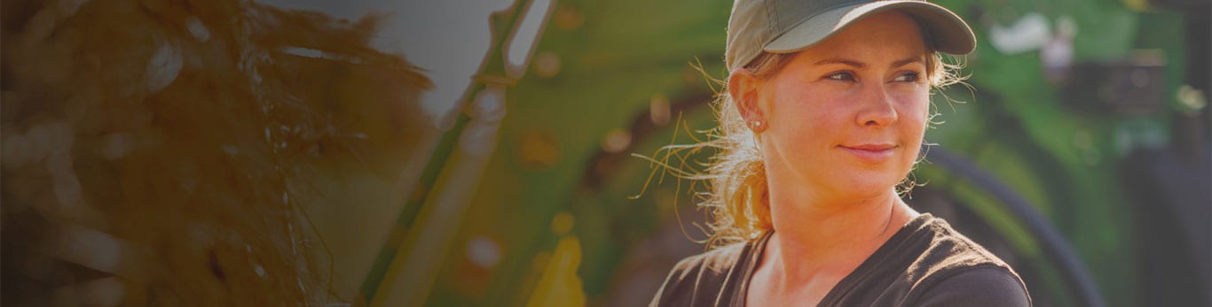 Person in a brown shirt and cap working with hay with a green tractor in the background