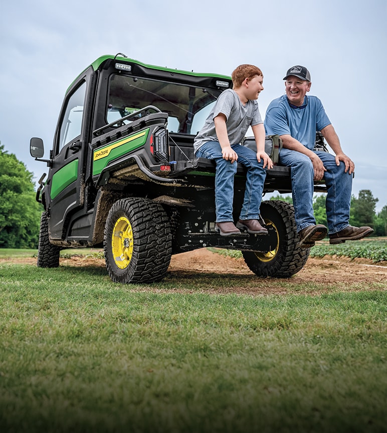 Man and boy sitting on the back of a John Deere Gator Utility vehicle on the edge of a forest