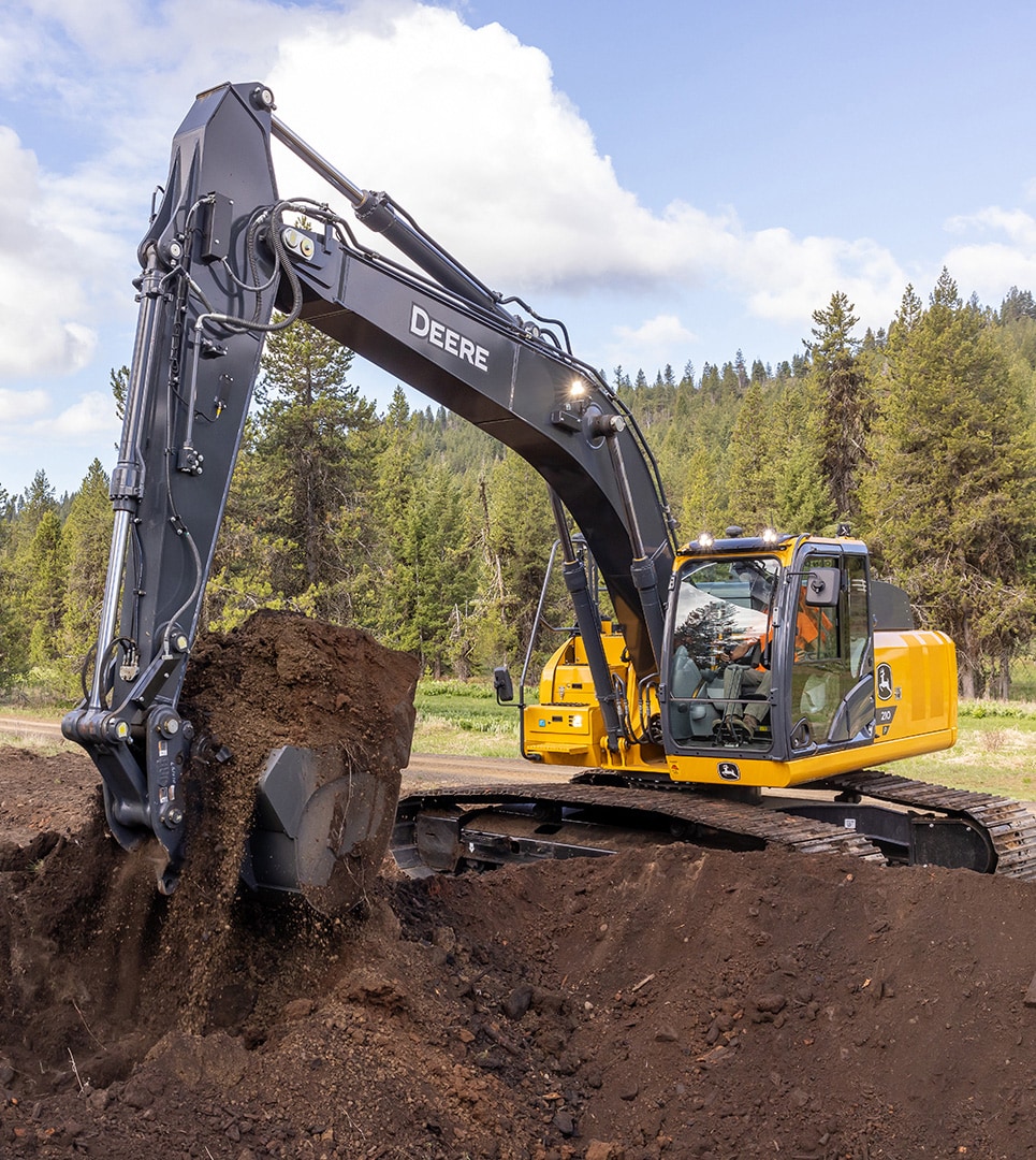 The John Deere 210P Excavator is digging a hole at a job site located in the woods