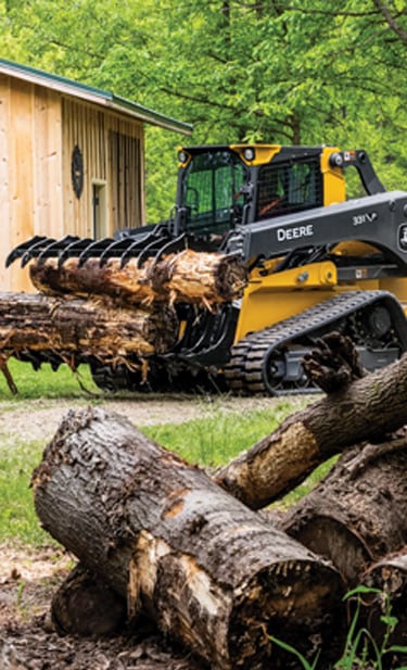 a 331 P-Tier CTL equipped with an RR84 Root Rake to move logs by a garage