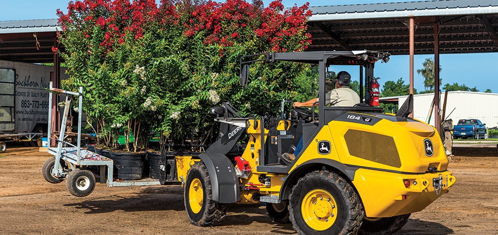 A John Deere 184 G-Tier Compact Wheel Loader with a pallet fork attachment transfers plants at a nursery.