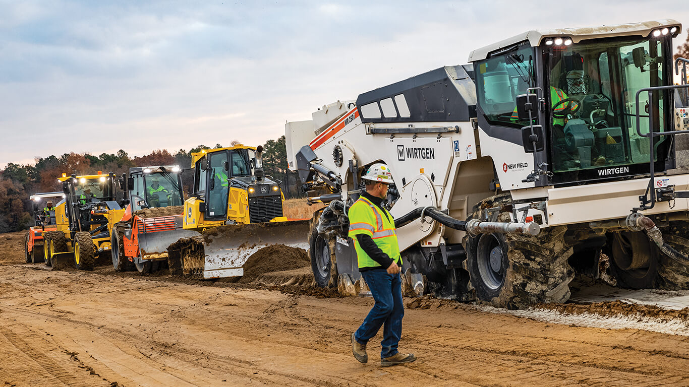 A person wearing construction gear stands on the dirt path of a jobsite looking back at John&nbsp;Deere and Wirtgen machines working together to build a road.