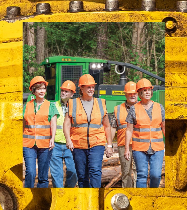 Group wearing orange safety vests and hard hats walking in front of green forestry machine.