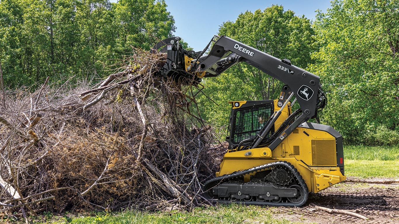 A John Deere compact track loader clearing a large pile of brush