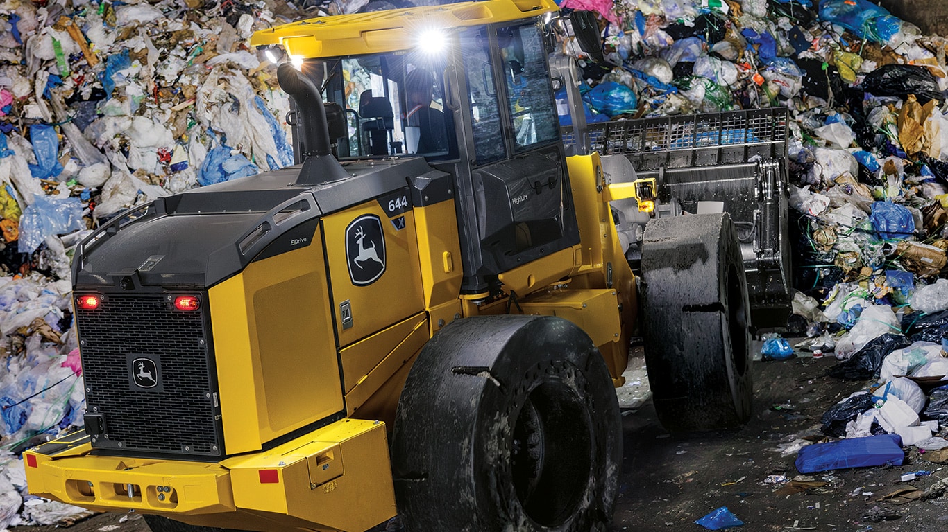 A John Deere 644 X-Tier Wheel Loader with a waste-moving bucket pushes trash into a pile.