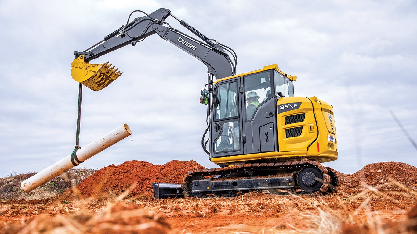 A yellow John Deere excavator uses its arm to lower a large, white pipe into the ground at a construction site in a dirt field.