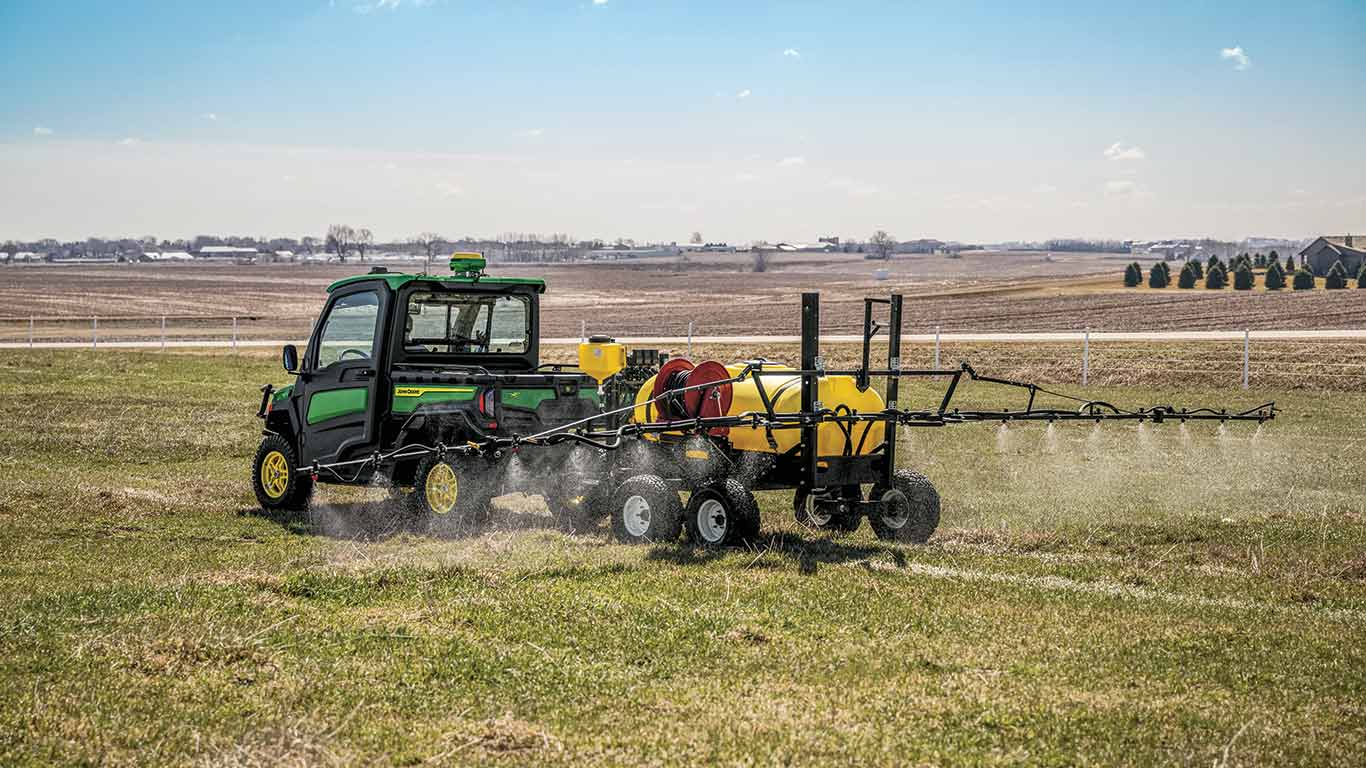 New John Deere Gator XUV 845R Signature Series Utility Vehicle with StarFire receiver in a field using a tank and sprayer boom rear attachment to apply chemical to the field on a bright sunny day with blue skies and minimal clouds