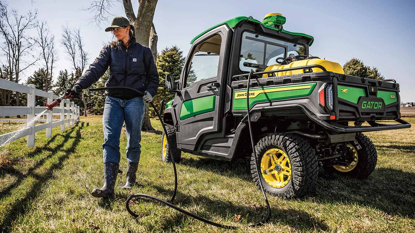 Operator is outside of a stationary new John Deere Gator XUV Signature Series Utility Vehicle with StarFire receiver to use a hose to spray ground around a white picket fence as the hose is attached to a 45-gallon yellow tank in the back of the Gator's large cargo box
