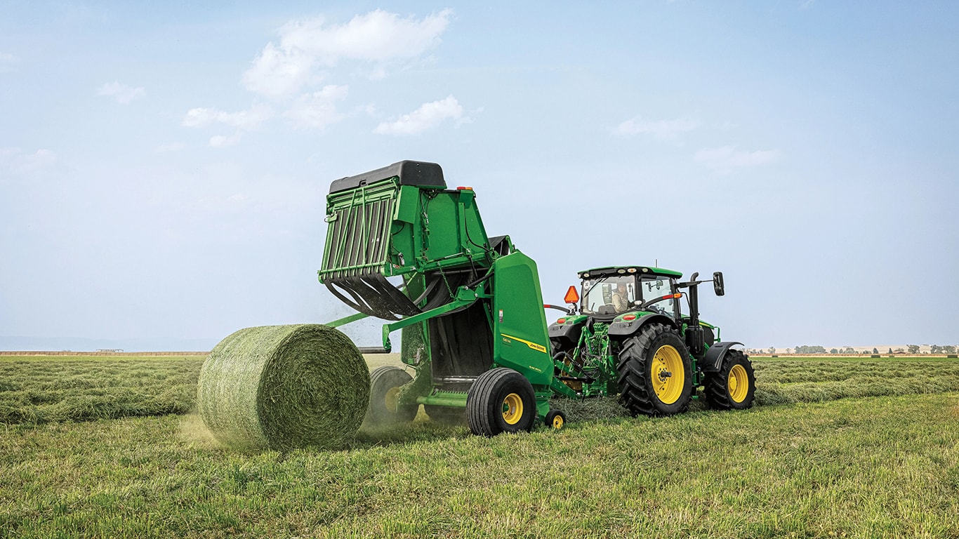 John&nbsp;Deere 6R Series Tractor pulling a John&nbsp;Deere 1 Series Round Baler in a hay field with a newly-produced round bale being ejected from the back of the baler as the baler's gate it flipped open.