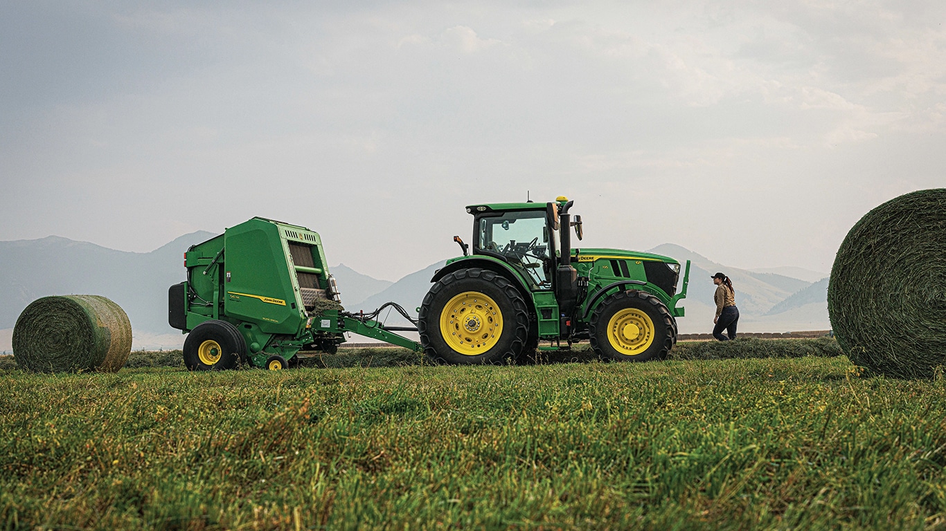 John&nbsp;Deere 6R Series Tractor pulling a John&nbsp;Deere 1 Series Round Baler in a hay field with a round bale behind the baler.