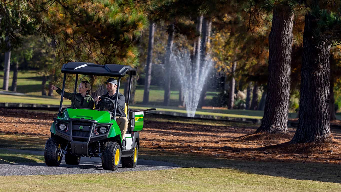 Man and woman riding in electric gator with water fountain behind them.