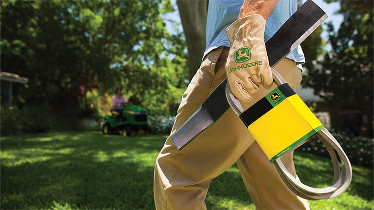 Close up, low angle image of a gloved hand holding mower blade and belt with John Deere branding with a residential lawn scene in the background.