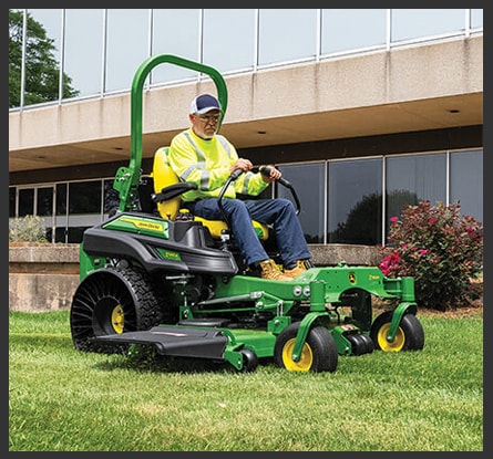 Person with a hi-vis vest, hat, and safety glasses operating a green zero-turn mower in front of a commercial building.