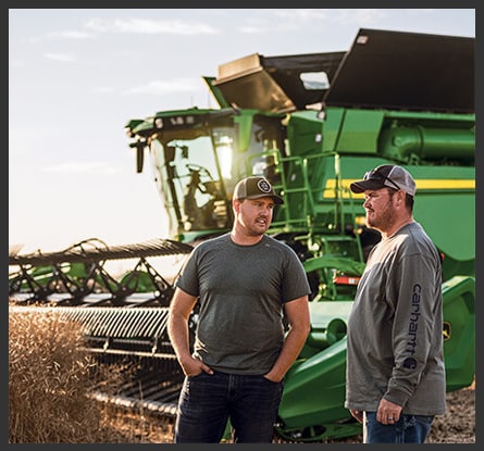 Two people talking to one another with a green combine and wheat field in the background.