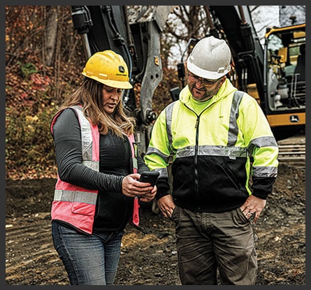 A person talking to another person while showing them their phone with an excavator, dirt, and trees in the background.