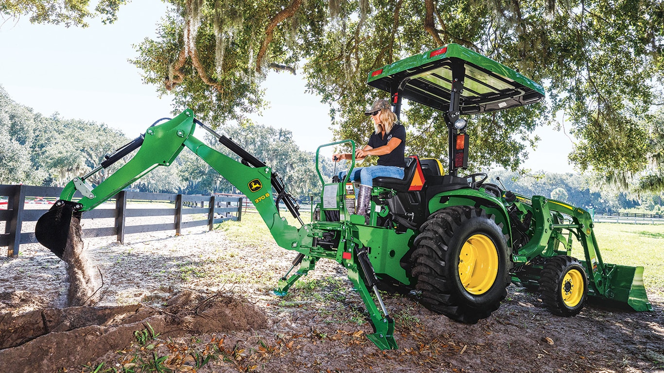 person with blonde hair and a ball cap operating a John Deere backhoe
