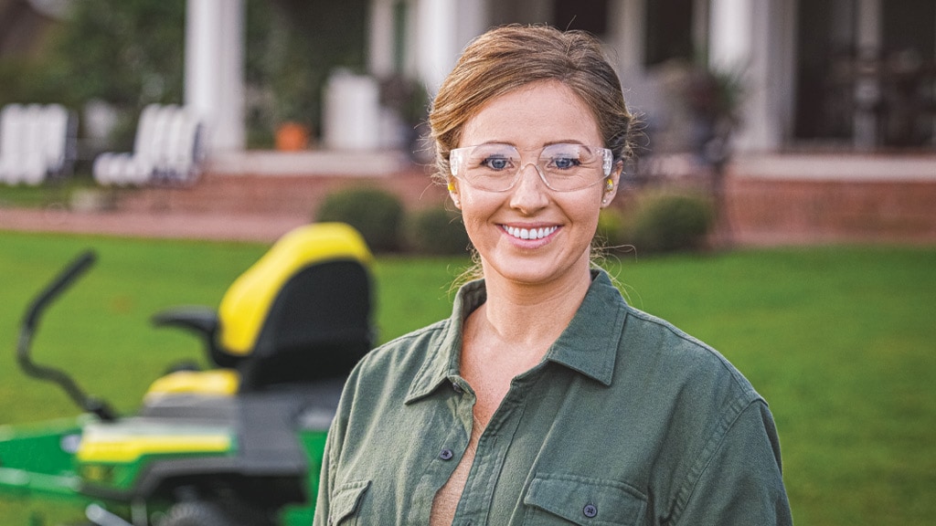 Person in a green shirt standing in front of a lawn mower with a house in the background.