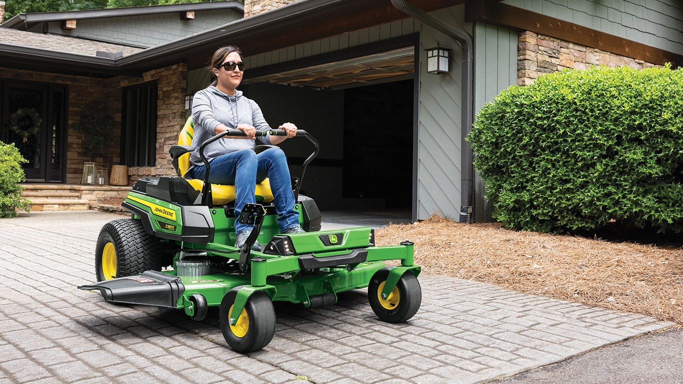 A woman with dark brown hair and a gray shirt and jeans on rides a green and yellow John Deere Z370R Electric lawn mower with safety glasses on.