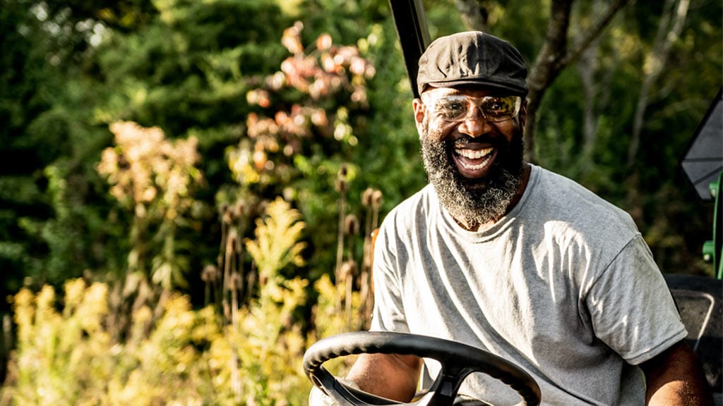 Man in a gray shirt driving a tractor outdoors with greenery in the background.