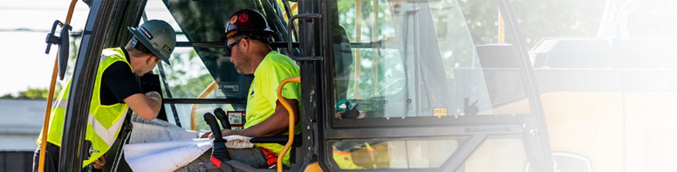 An excavator test pilot sits in a cab with another test pilot looking in at a diagram