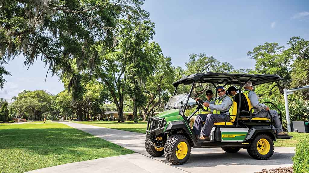 A green Gator carrying four passengers in work vests being driven down pavement.