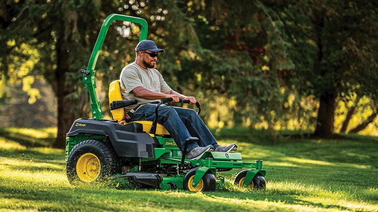 A person with sunglasses and a hat cuts grass with a green zero-turn mower in front of a line of trees. (11th thumbnail)