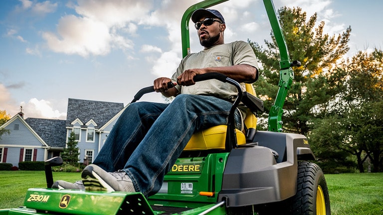 Close-up of a green zero-turn mower being driven with with a large lawn and blue residence in the background. (8th thumbnail)