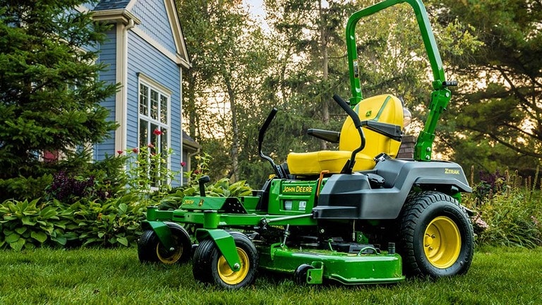 A green zero-turn mower rests on a well-kept lawn near a blue residence and garden. (7th thumbnail)