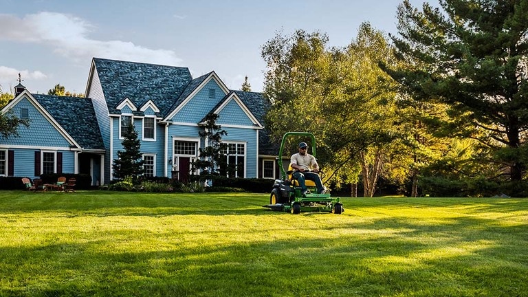 A far-off shot of a green zero-turn mower cutting a spacious lawn behind a large blue house. (9th thumbnail)
