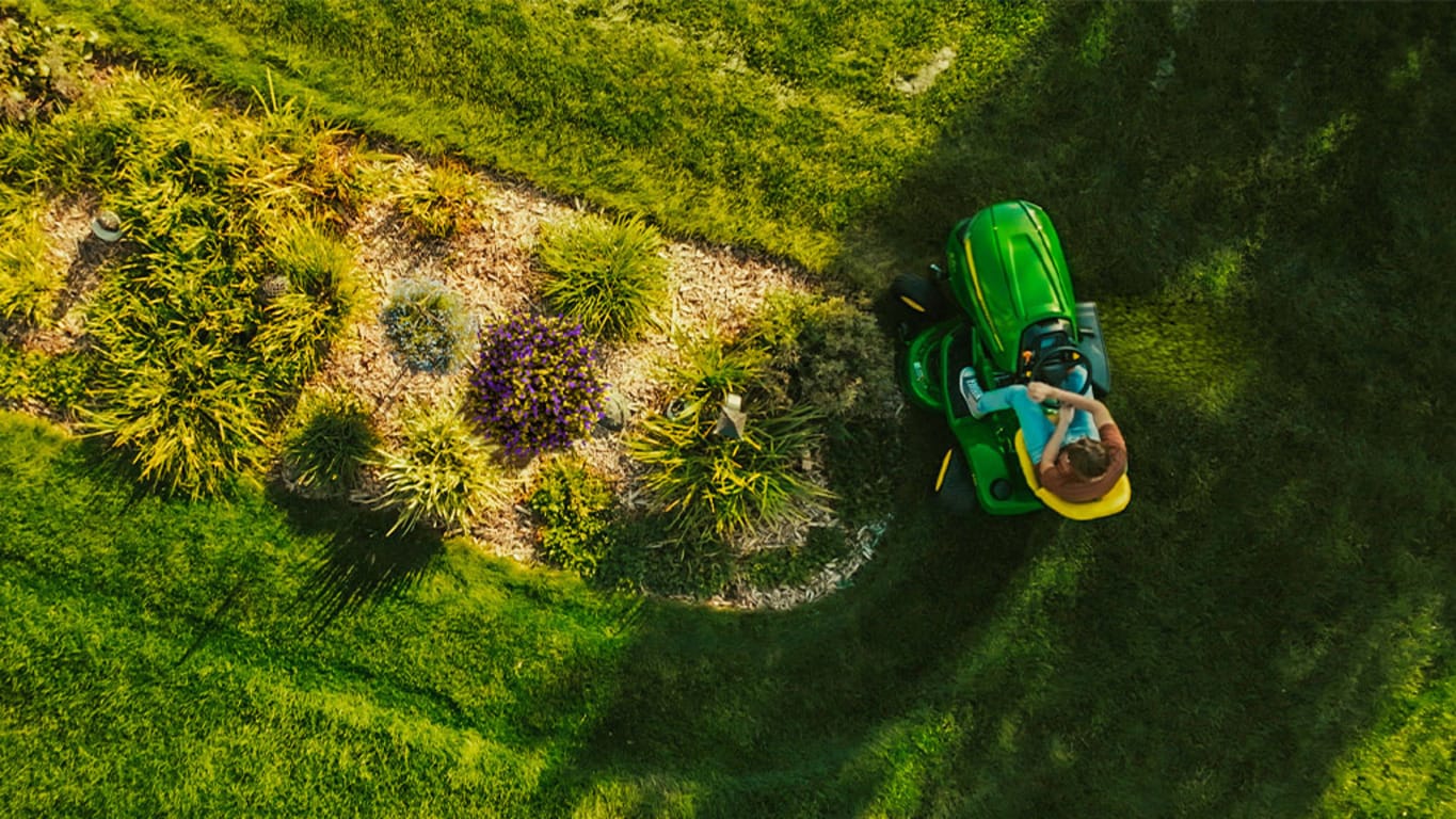 Top-down view of a green riding mower cutting grass around an oval-shaped garden patch.