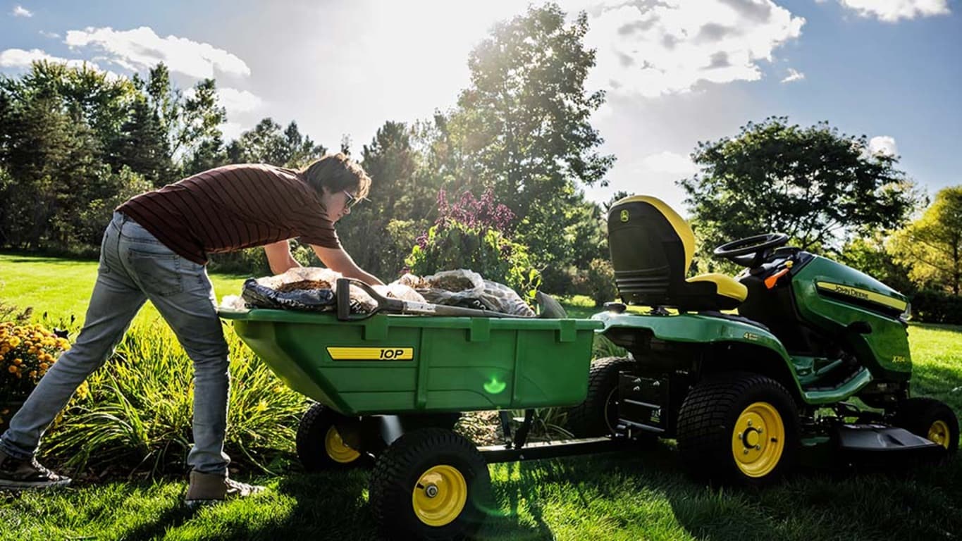 A person placing bags in a green polycart attached to the back of a riding mower.