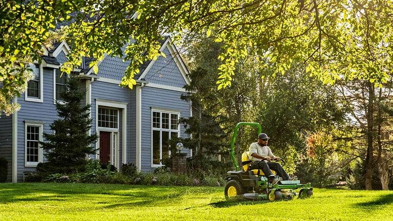 An under-tree shot of green zero-turn mowing along a treeline near a residence on a bright sunny day. (10th thumbnail)