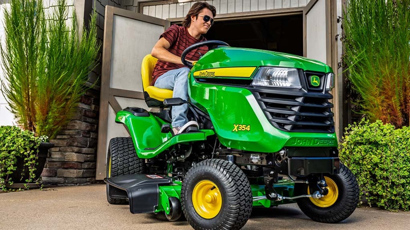 A person with sunglasses drives a green riding mower out of open shed doors.