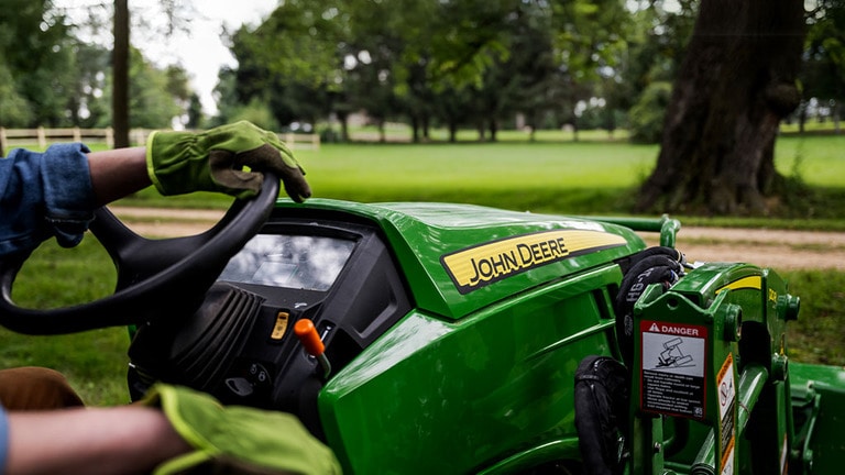 Close up of the John Deere logo on the front hood of a compact tractor while a gloved hand rest on the steering wheel. (11th thumbnail)