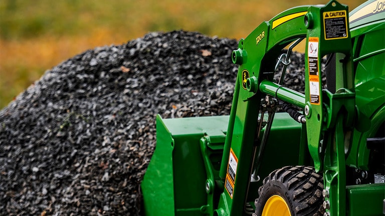 A green front loader attached to a compact tractor digs into a pile of gravel ready to lift it up. (12th thumbnail)