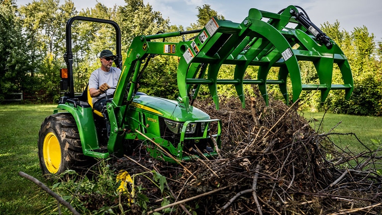 A debris grapple attached to a green compact tractor is open about to grab a pile of branches. (2nd thumbnail)