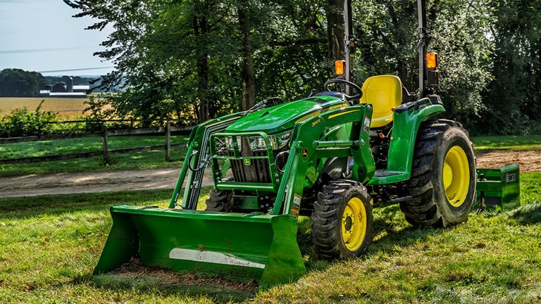 A person approaches a green compact tractor resting in the sun with a loader and box blade attached to it. (3rd thumbnail)