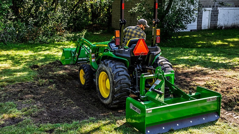 A front-loader and box blade attached to a green compact tractor scrapes off the top soil and grass to create a dirt patch. (7th thumbnail)