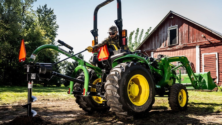 A post-hole digger on the back of a green compact tractor is creating a hole near a red barn. (6th thumbnail)