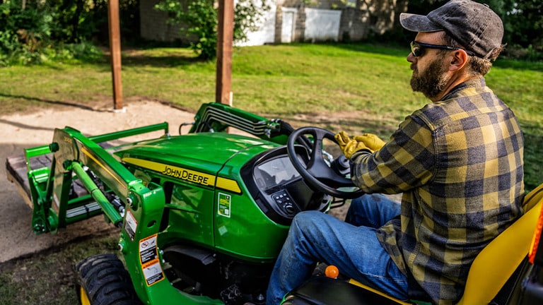 A pallet of pavers is carried to a cleared dirt patch by a green compact tractor with a pallet fork attachment. (5th thumbnail)