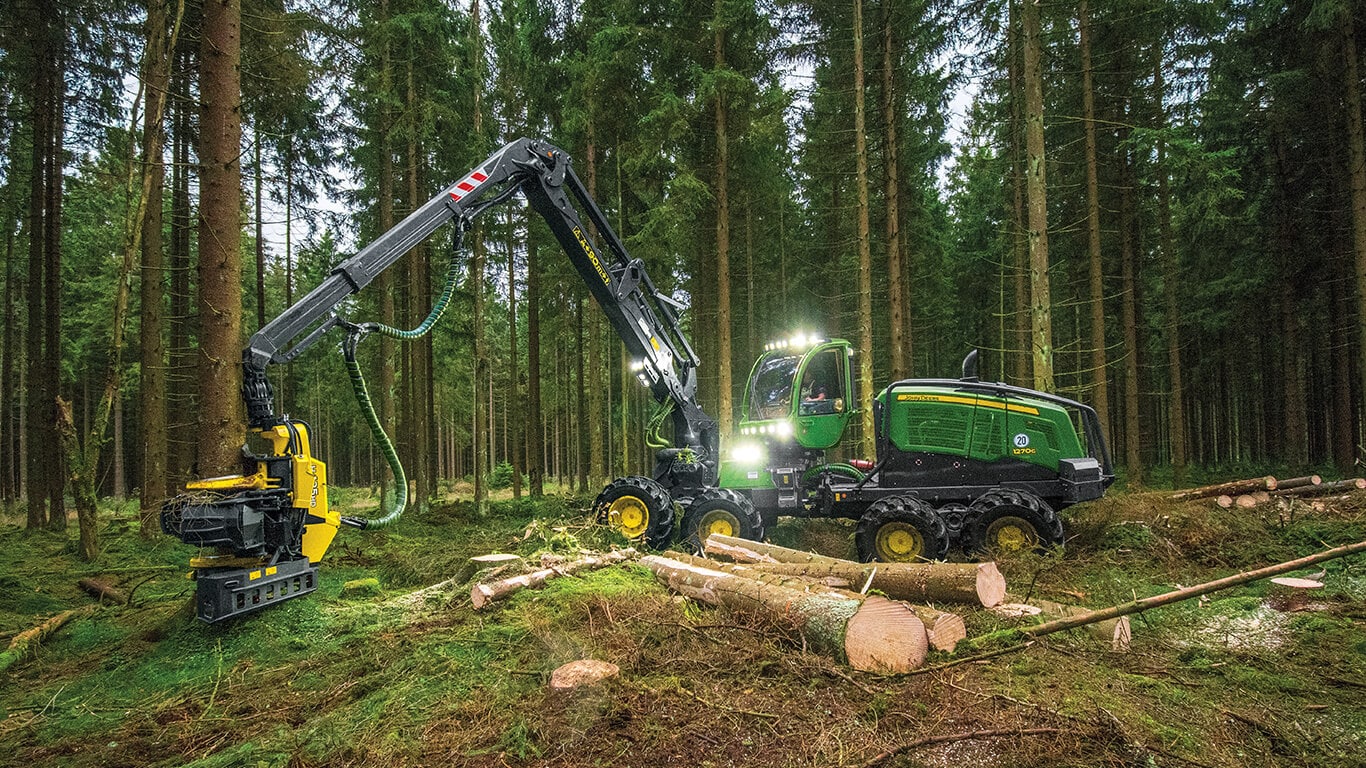 John Deere wheeled harvester working in wooded area with lights on harvesting trees.