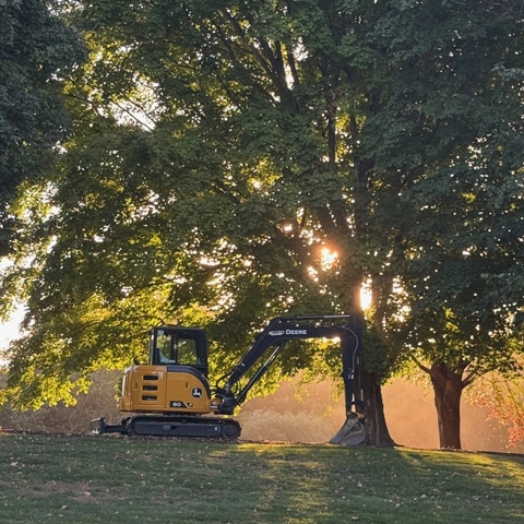 A John Deere 60 P-Tier Compact Excavator is parked in the shadow of trees, sitting on the ridge of a hill.