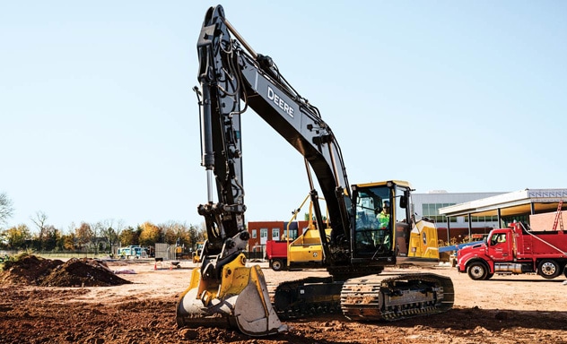 Tracked excavator digging soil at an active construction site with trucks and buildings nearby.