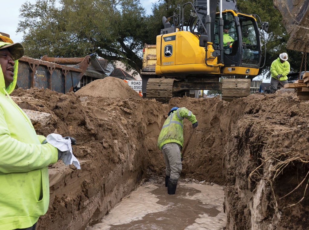 A worker uses a shovel inside a large trench surrounded by large construction equipment