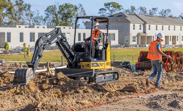 John Deere 26P-Tier Compact Excavator digging into the ground with building structures in the background